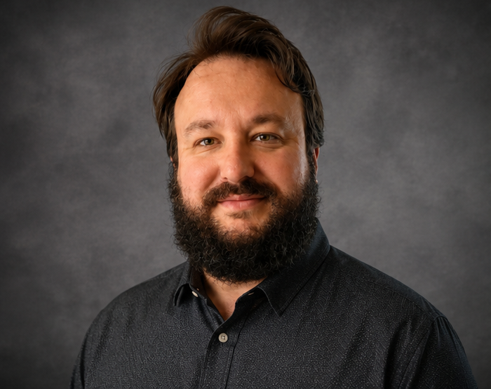 Bearded man with dark hair wearing a black button-up shirt against a gray backdrop.