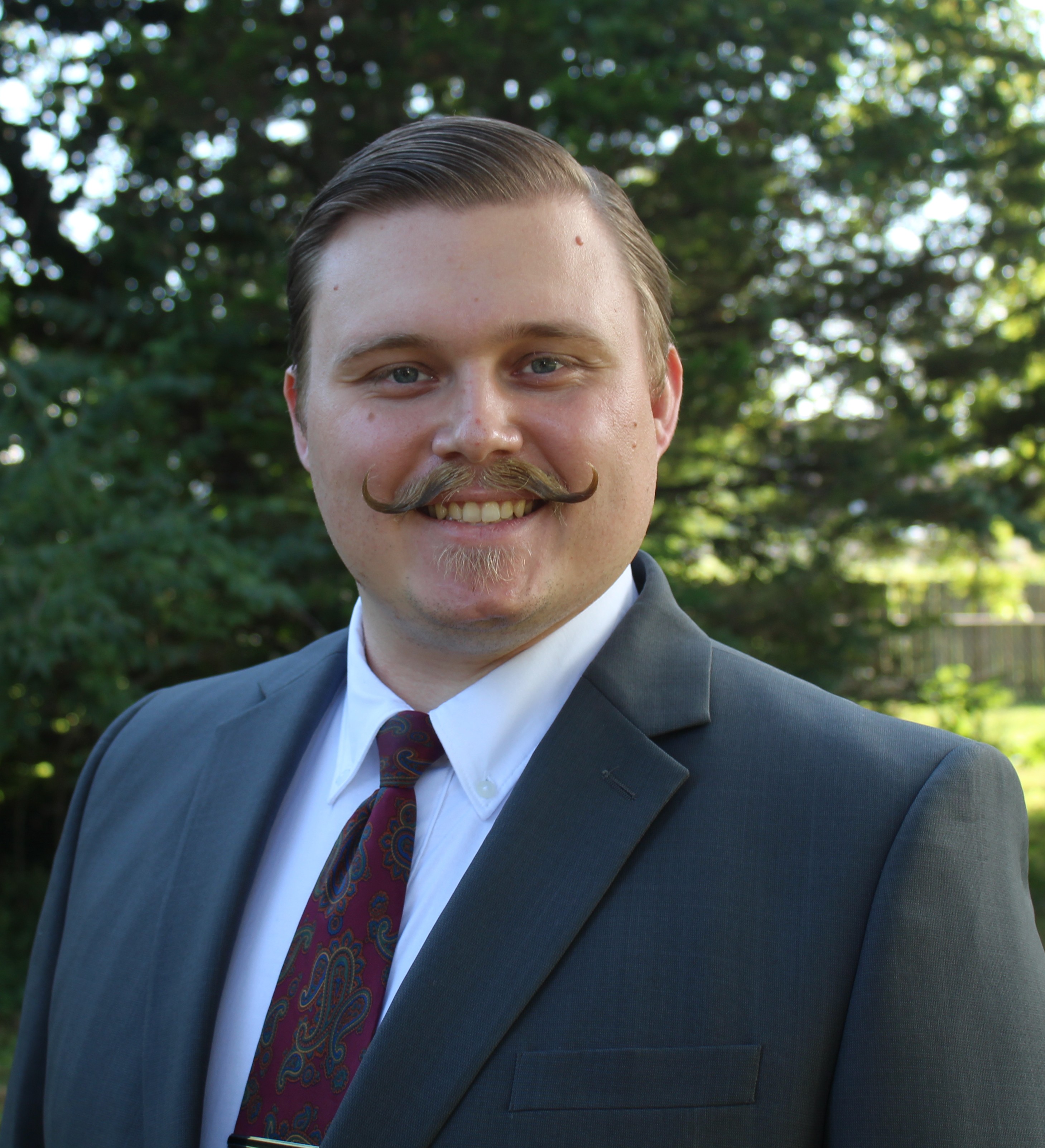 Smiling man with styled mustache wearing a dark suit, white shirt, and patterned tie outdoors.
