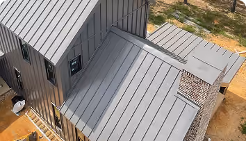 Aerial view of a modern house with a grey metal roof, brick chimney, and surrounding dirt ground.