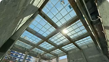 Sunlight shines through a metal grid roof structure with glass panels over a concrete building.