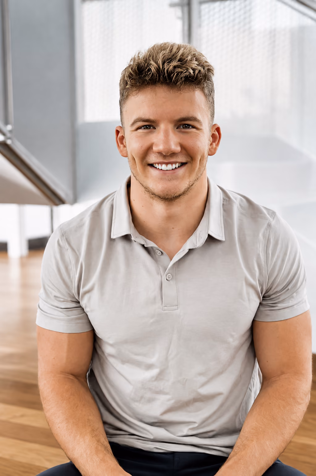 Smiling young man with curly hair wearing a polo shirt, seated indoors with a bright background.
