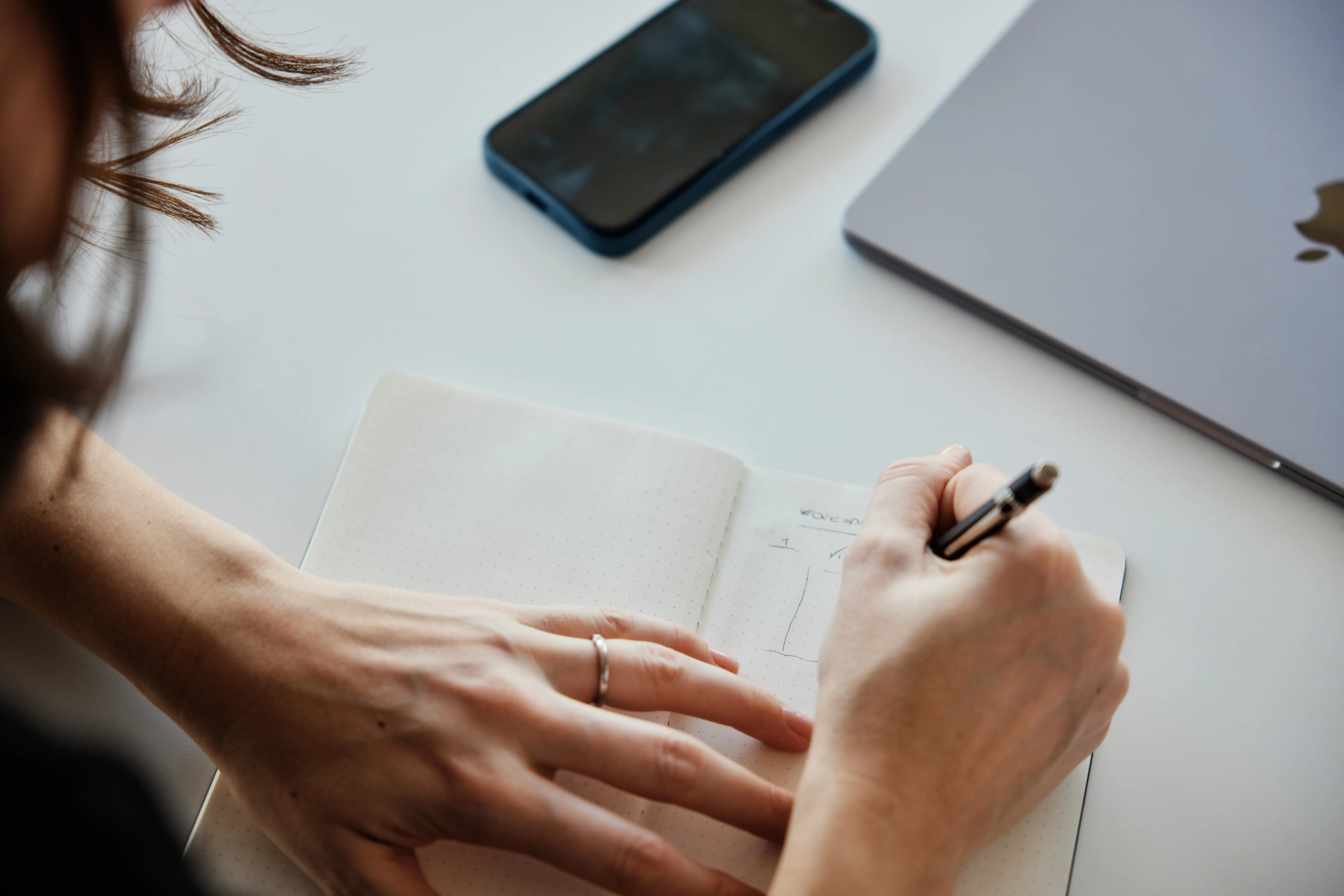 Hand macht Notizen in einem Notizbuch, daneben ein Smartphone