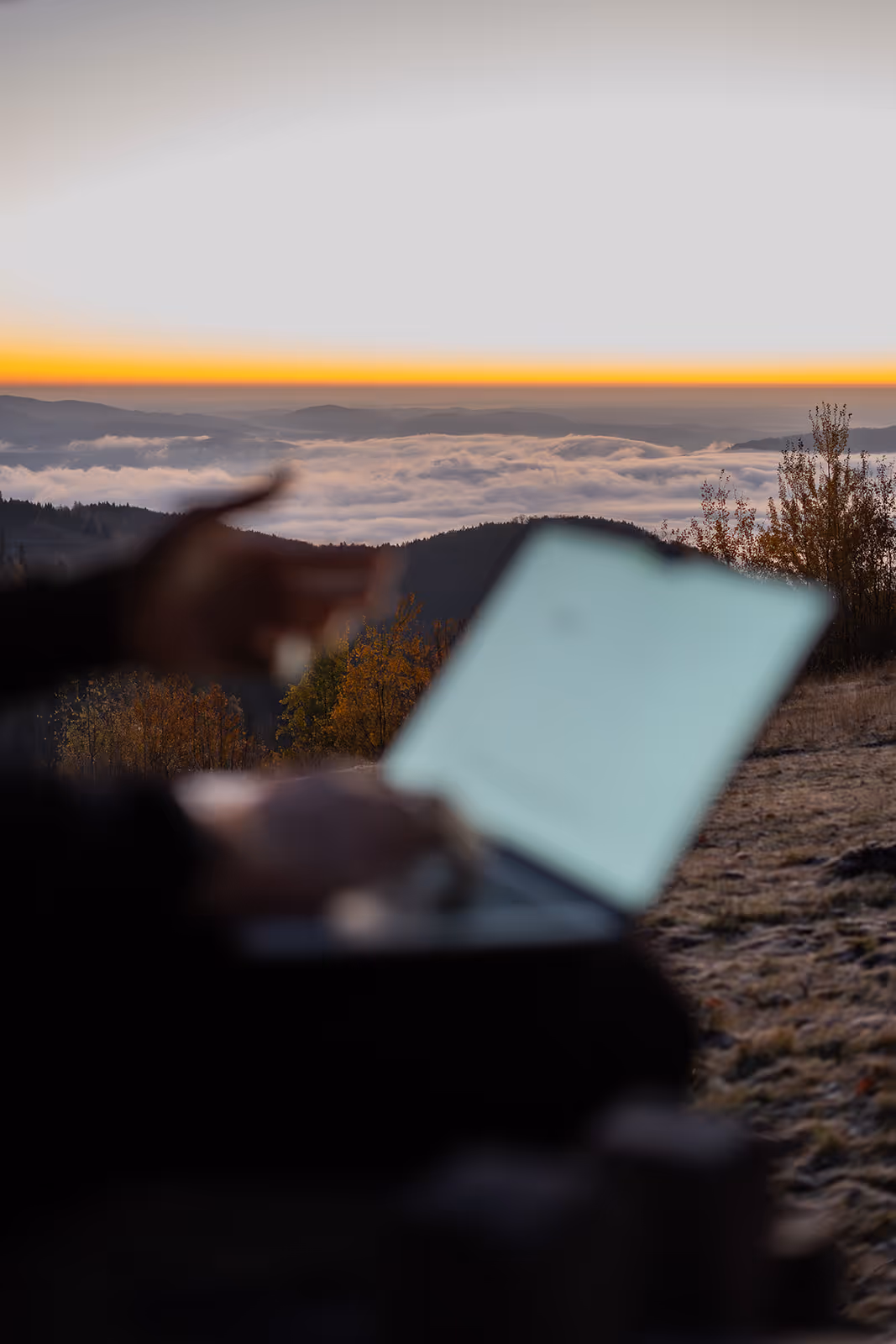 Person arbeitet mit Laptop im Freien vor einem Nebelmeer bei Sonnenaufgang in den Bergen.