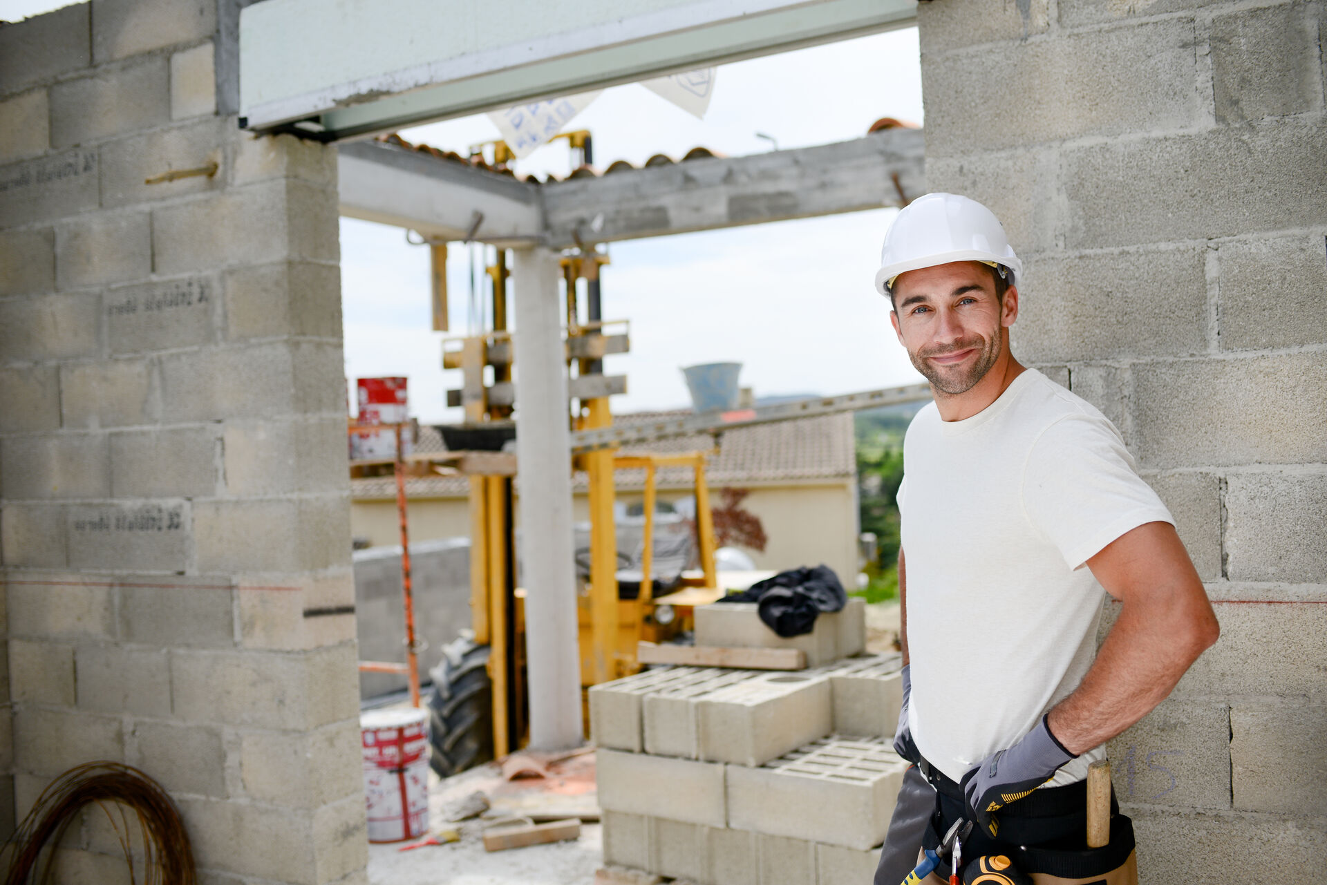 A construction worker wearing a hard hat and tool belt stands inside a partially built concrete-block structure.