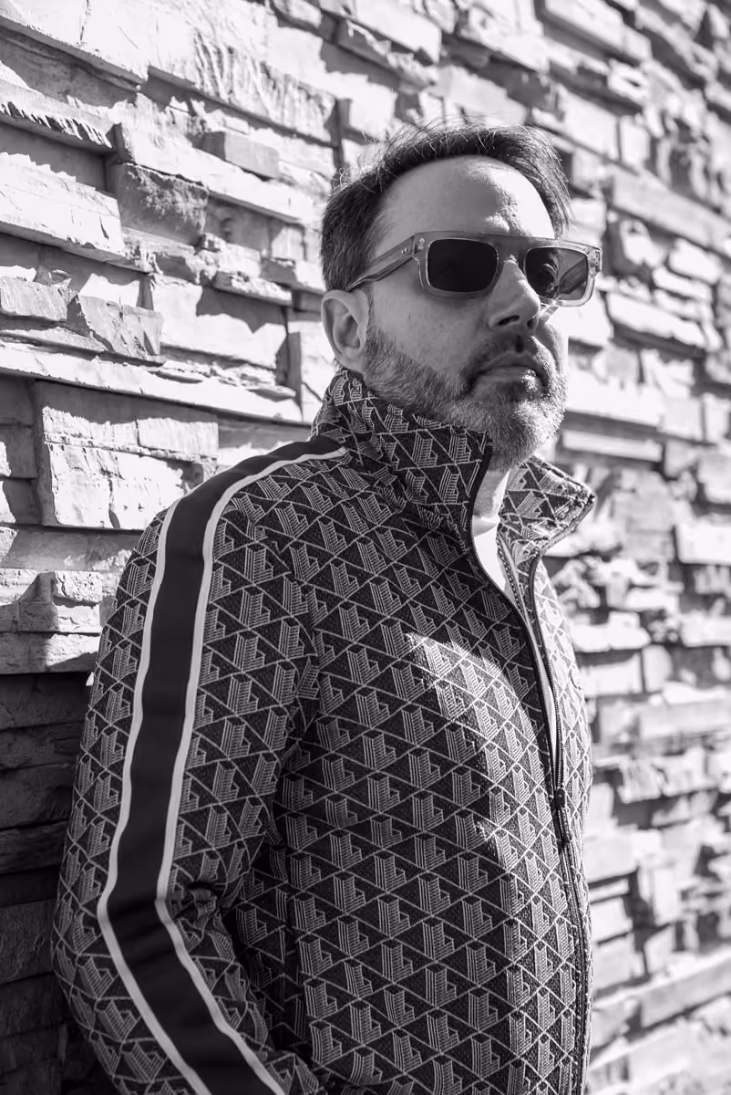 Man with short hair and beard wearing sunglasses and patterned jacket stands against textured stone wall.