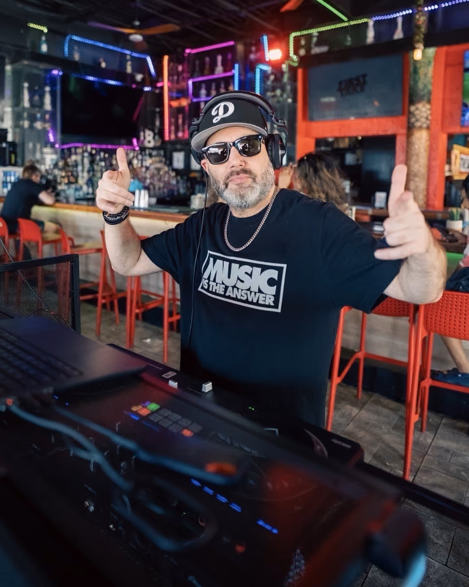 Man wearing sunglasses and headphones DJing at a bar with neon lights and red stools in the background.