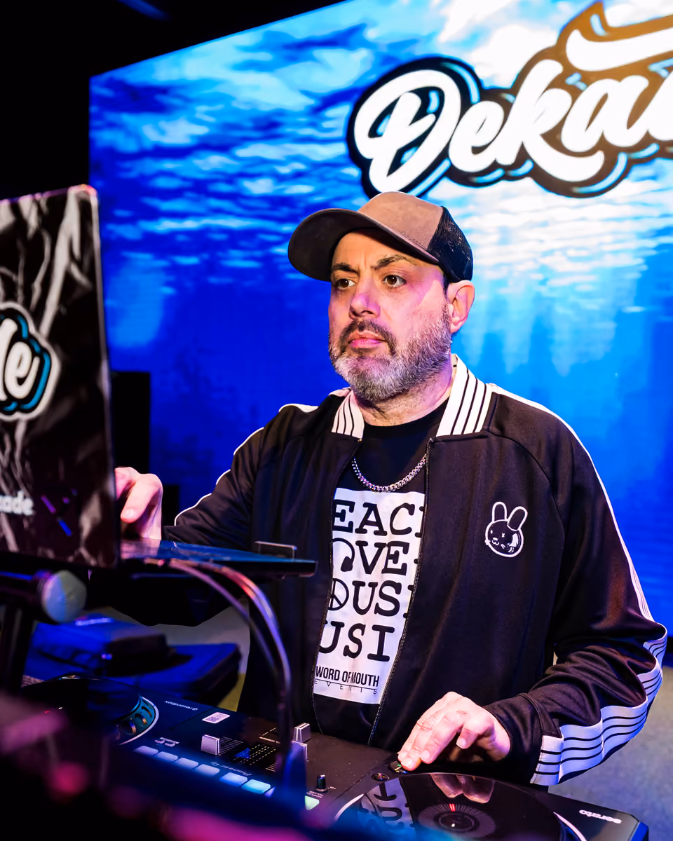 DJ wearing a cap and jacket performing with a mixer, with a Dekade logo in the blue background.