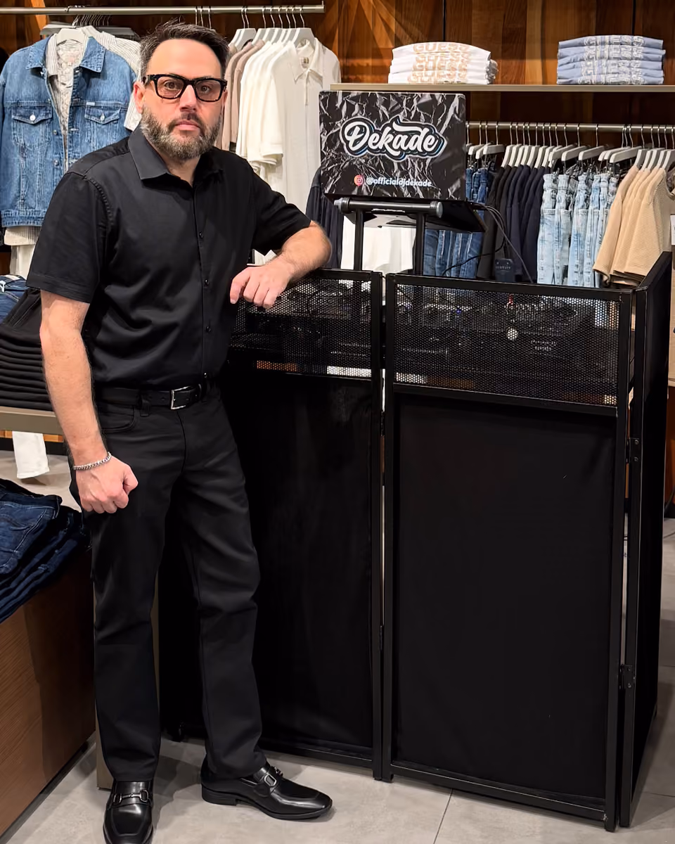 Man in black outfit and glasses standing next to DJ booth with Dekade logo inside a clothing store.