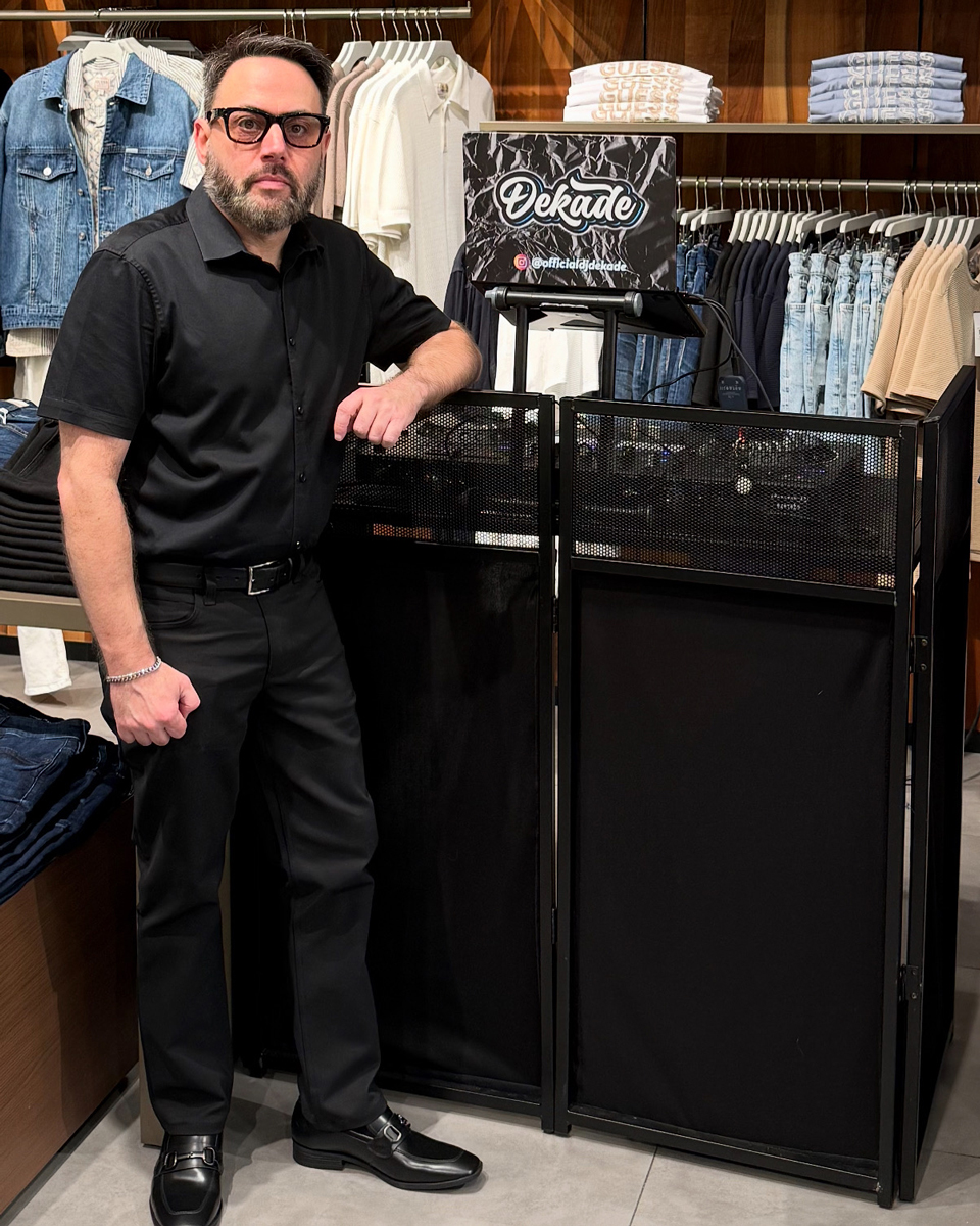 Man in black outfit and glasses standing next to DJ booth with Dekade logo inside a clothing store.