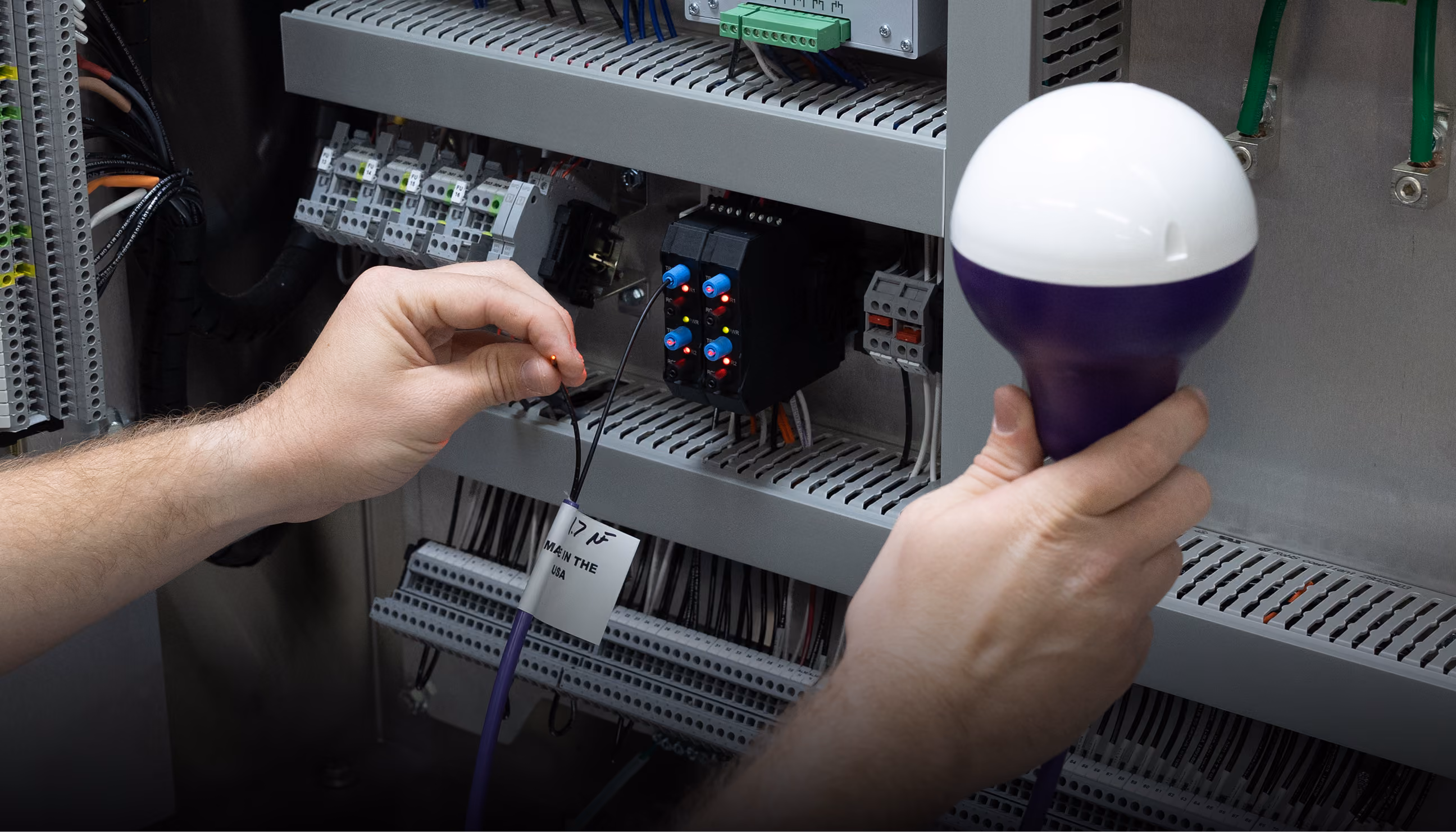 Person holding a cable with a connector and a device in front of an electrical control panel.