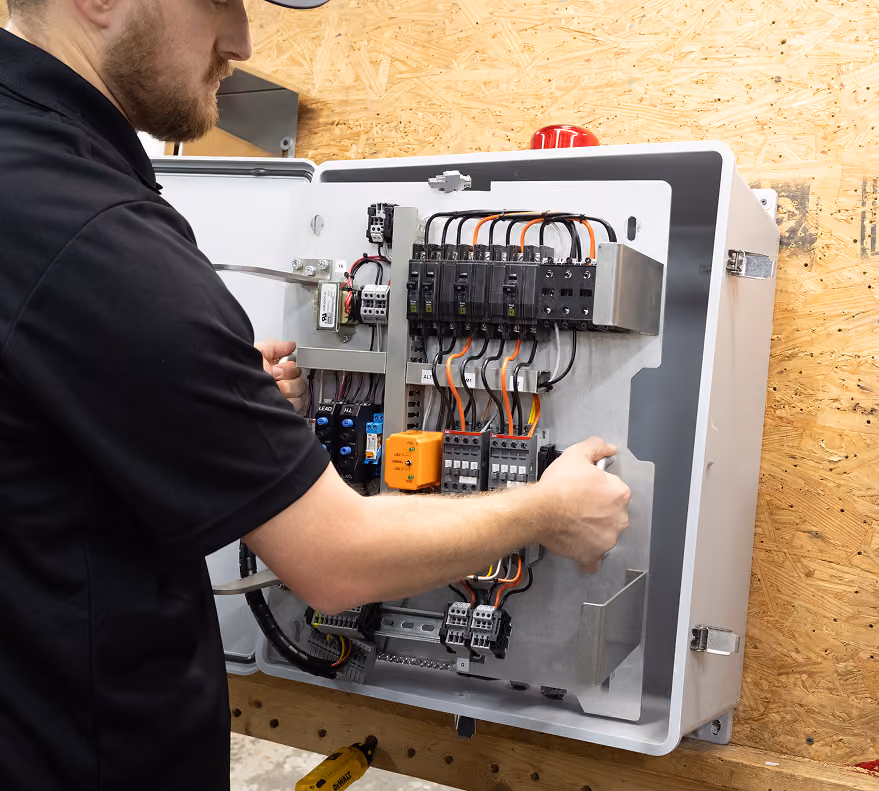 Man in a black shirt working on an open electrical control panel with wires and switches.