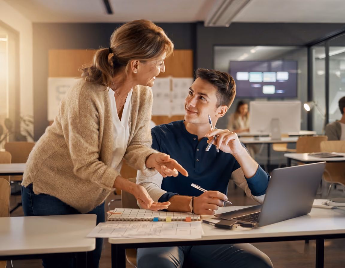 Woman and young man discussing work at a desk with laptop and documents in an office.