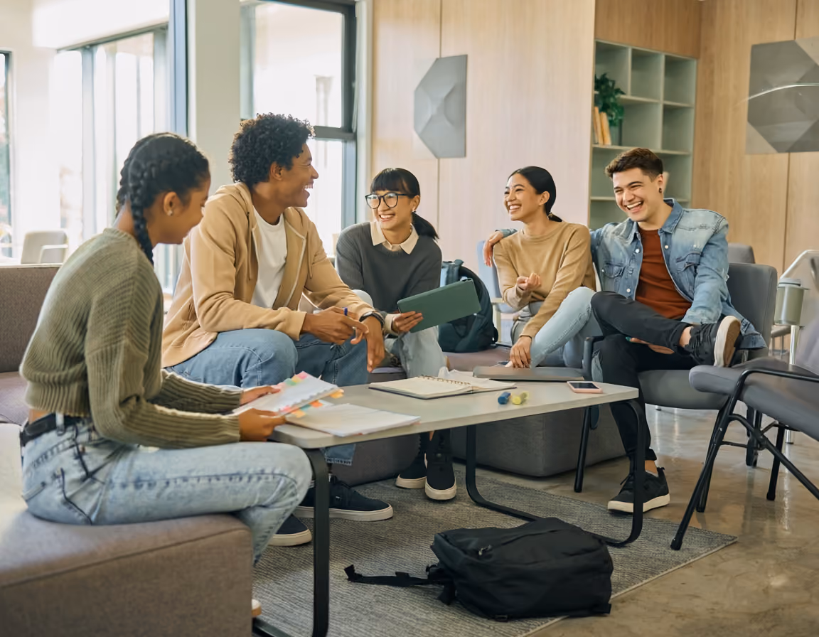 Group of five young adults sitting in a lounge area studying and chatting with notebooks and a tablet.