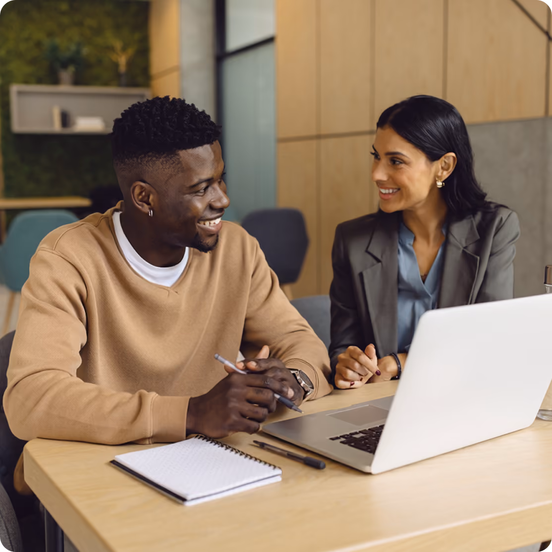 Two colleagues smiling and discussing work at a table with a laptop and notebook.