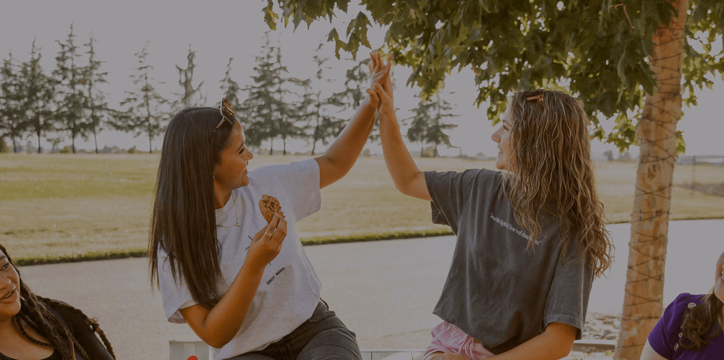 Two young women outdoors high-fiving, one holding a cookie, with trees in the background.