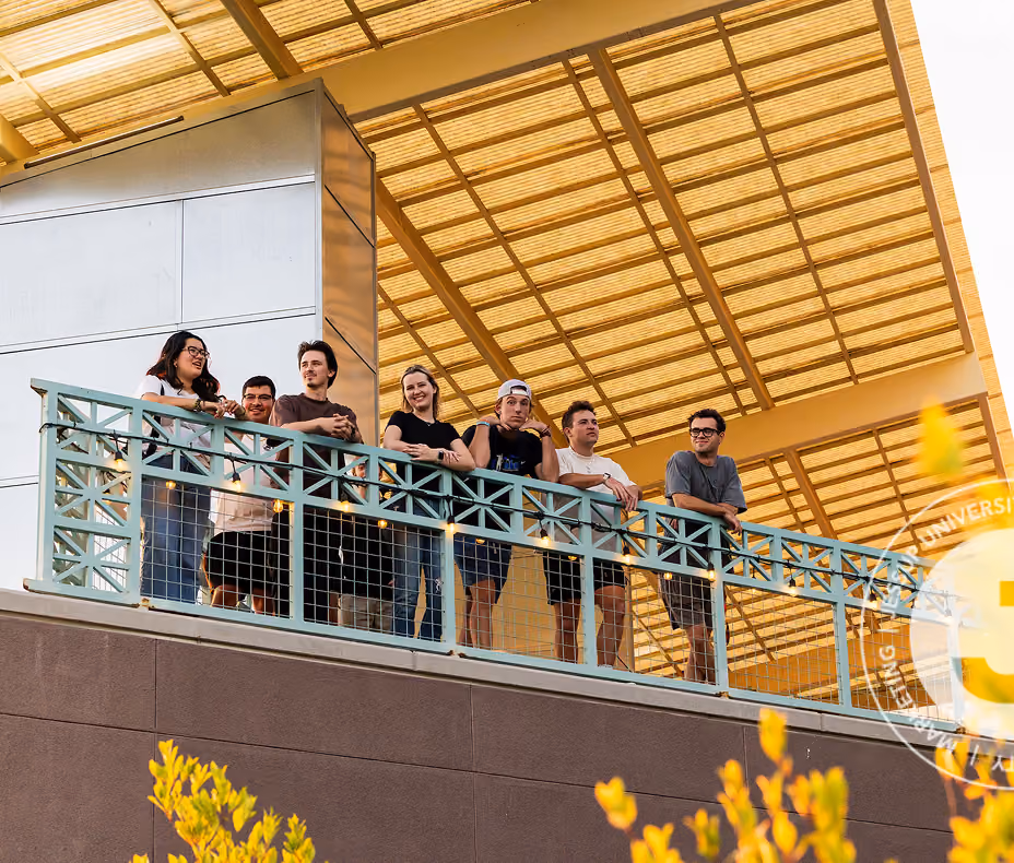 Seven young people standing on a balcony under a large grid-like roof, smiling and looking out.
