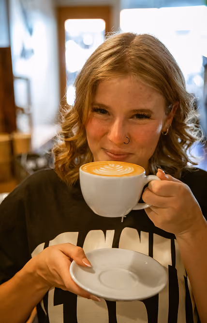 Smiling woman with curly hair holding a cup of latte with latte art in a cafe.