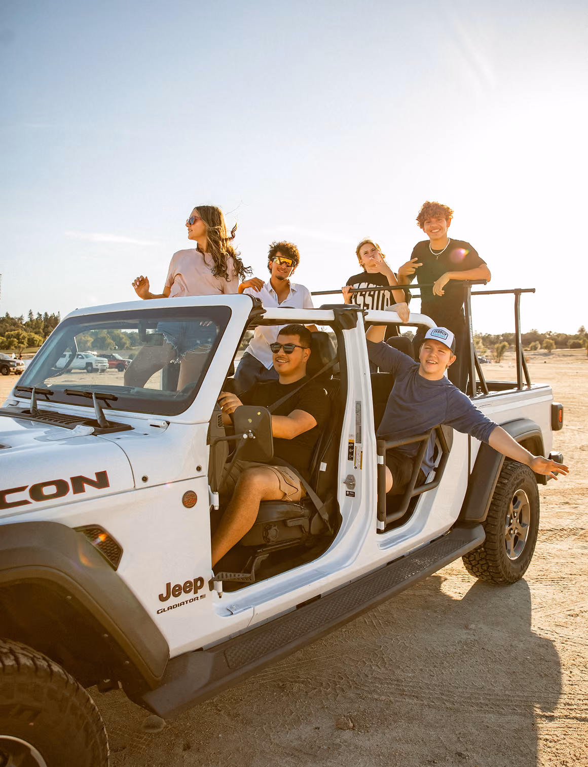 Group of six young people enjoying a sunny day in a white Jeep Gladiator with open doors.