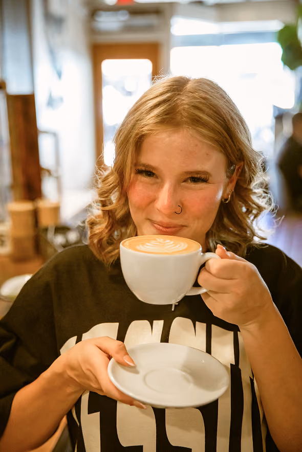 Young woman with light brown hair holding a coffee cup with latte art and smiling.