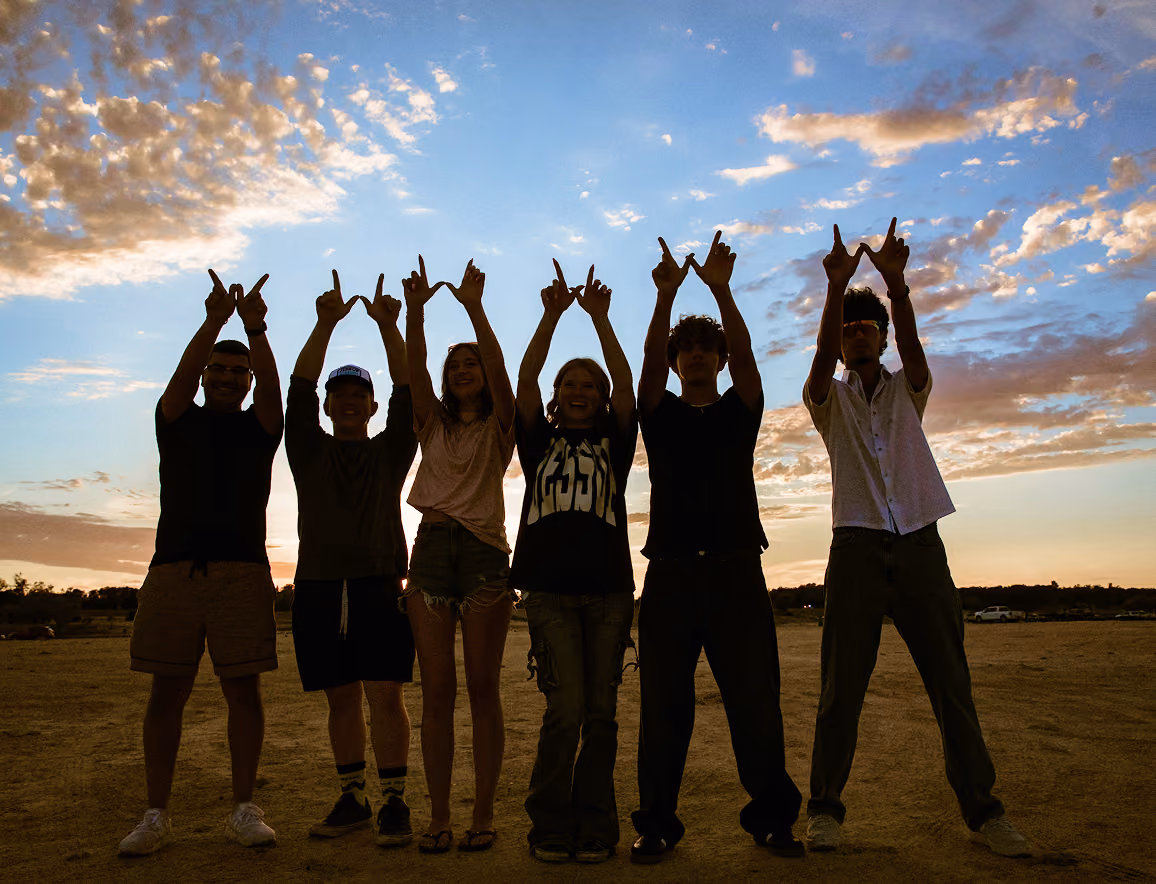 Six people standing outdoors at sunset with arms raised forming 'W' shapes with their fingers.