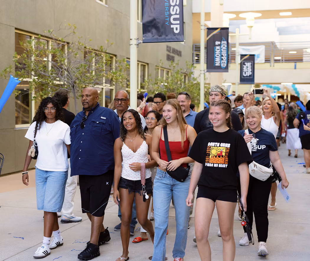 A diverse group of smiling people walking through a university campus courtyard with banners overhead.
