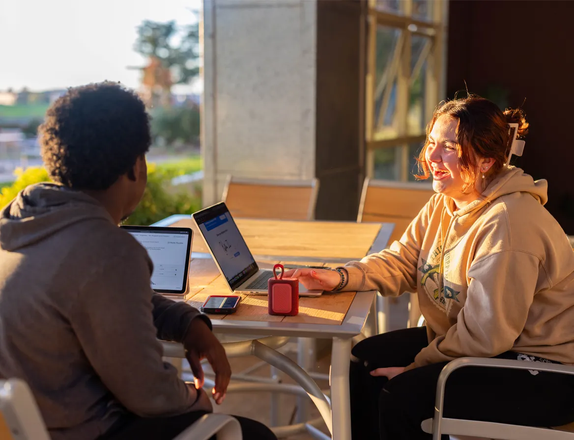 Two people sitting at a table outdoors, working on laptops and smiling in sunlight.