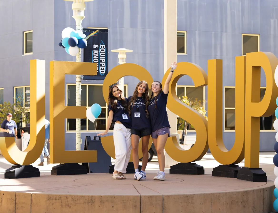 Three young women posing happily in front of large gold letters spelling JESUS UP.