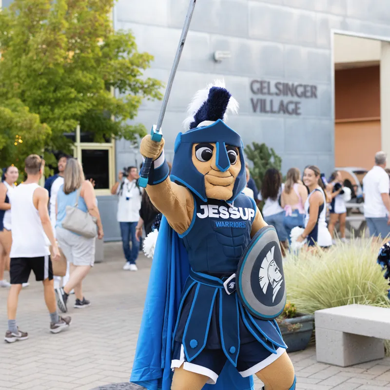 Jesup Warriors mascot in blue armor holding a sword and shield outdoors near a building sign reading Gelsinger Village.