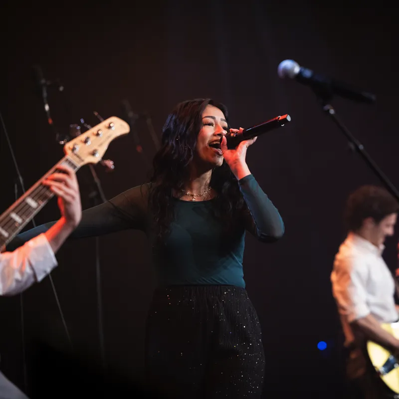 Female singer passionately performing on stage with musicians playing guitars.