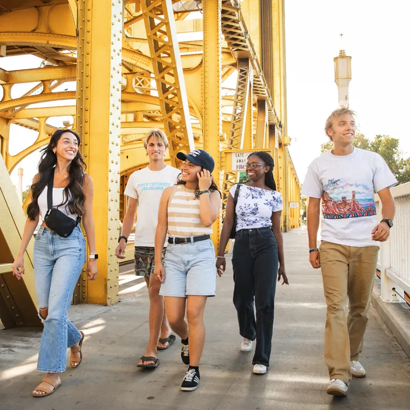 Five diverse young adults casually walking and smiling on a bright yellow steel bridge in daylight.