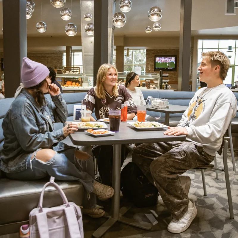 Three young adults sitting at a table eating food and talking in a modern cafeteria.