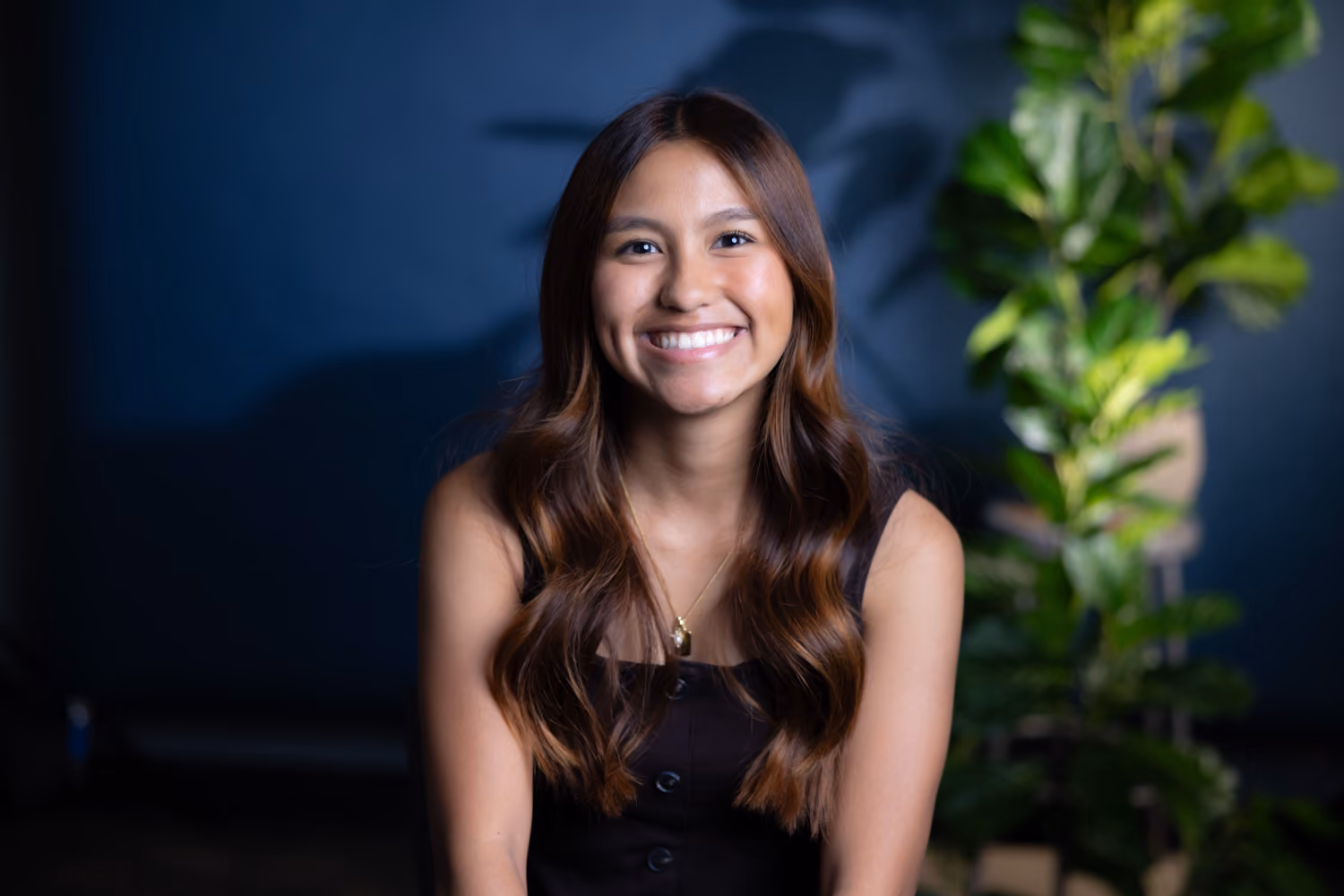 Smiling young woman with long wavy hair wearing a black sleeveless top sitting indoors near a green plant.