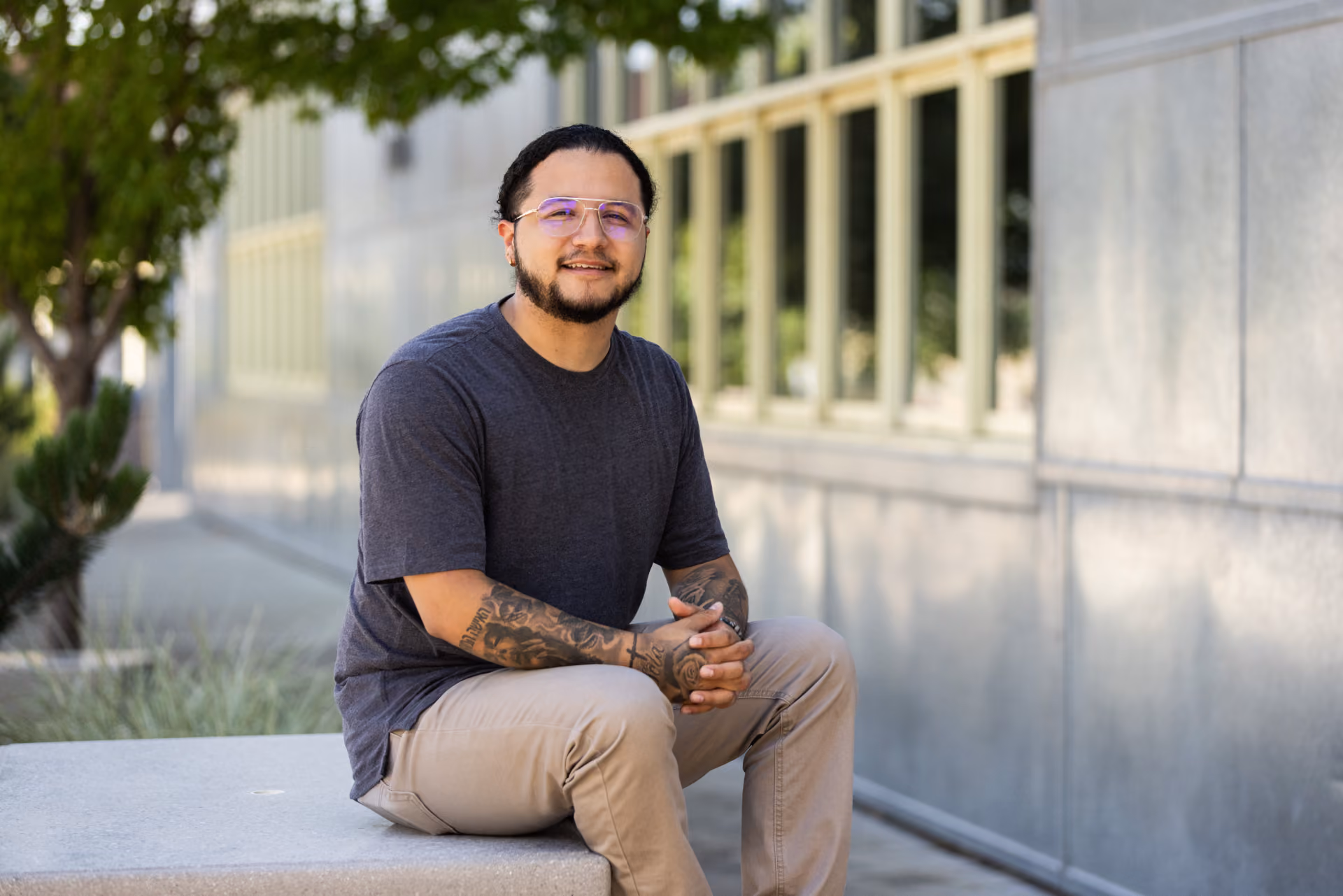 Man with tattoos wearing glasses and a gray shirt sitting on a concrete bench outside near a building.