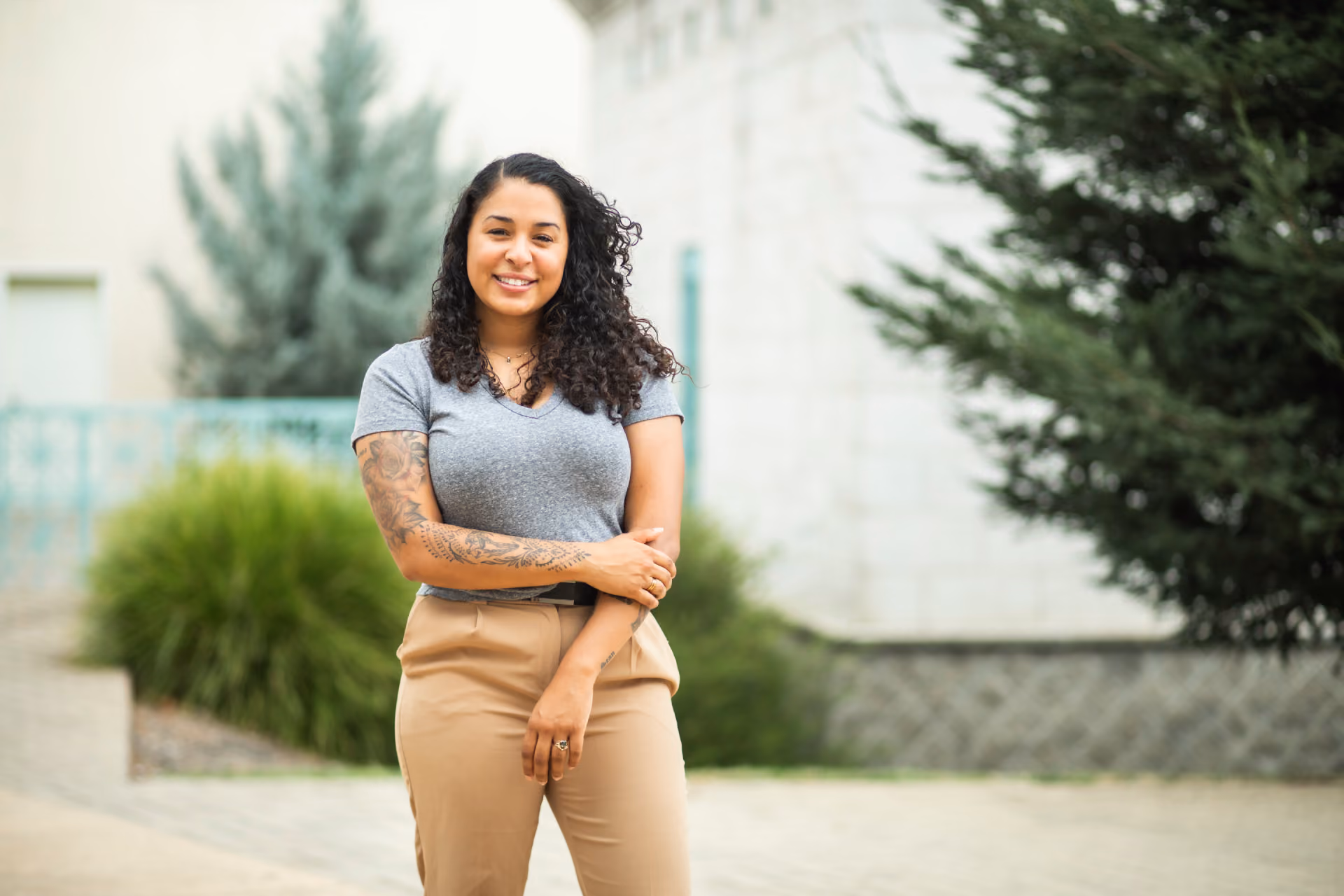 Smiling woman with curly hair and sleeve tattoo, wearing gray shirt and tan pants outdoors.