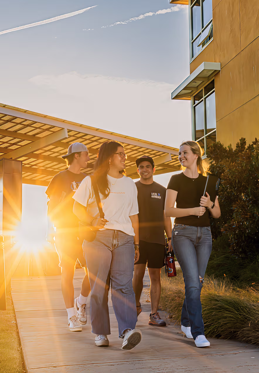 Four young adults walking and talking outside near a building at sunset, carrying books and a water bottle.