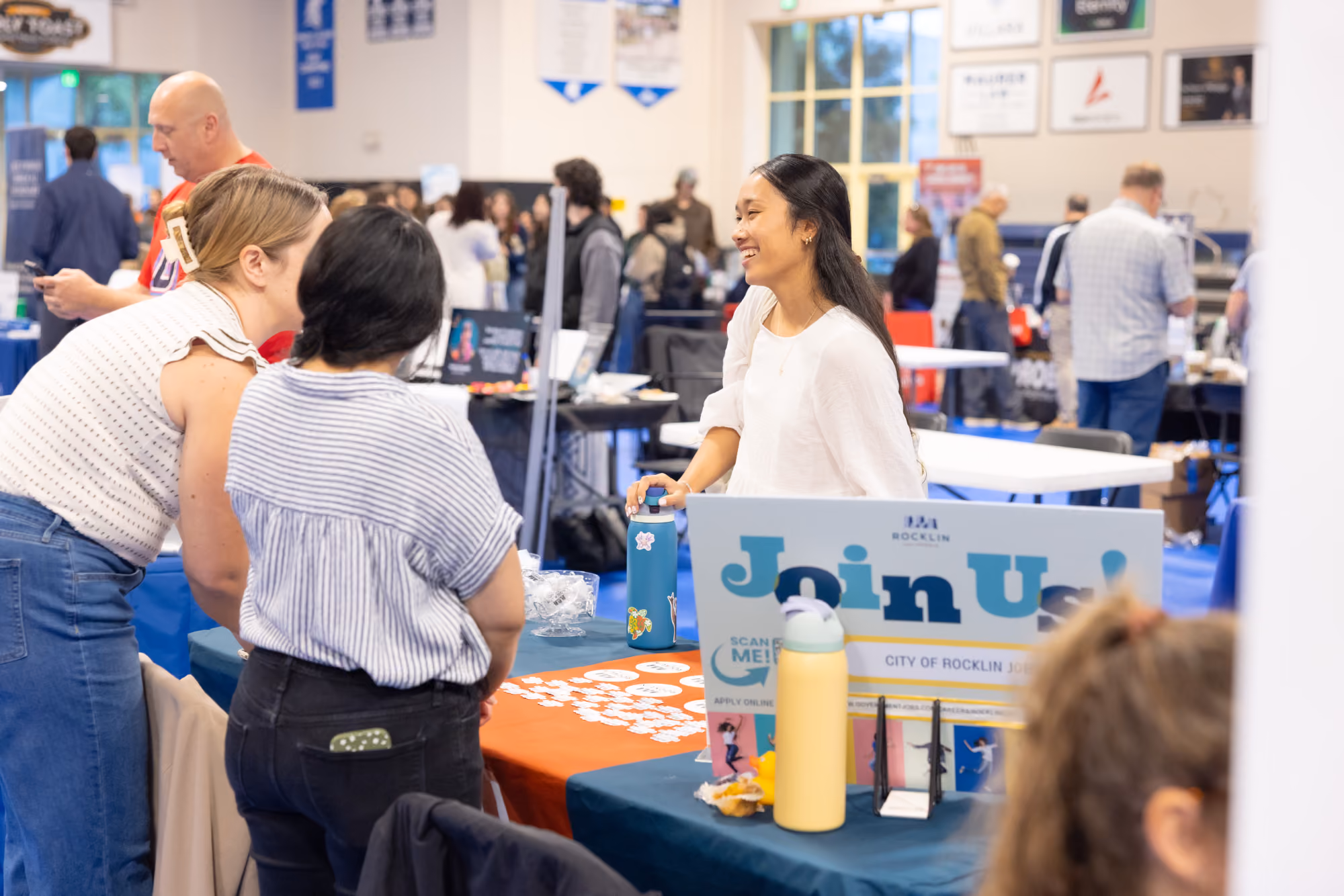A smiling woman talks with visitors at a City of Rocklin jobs booth during an indoor event.