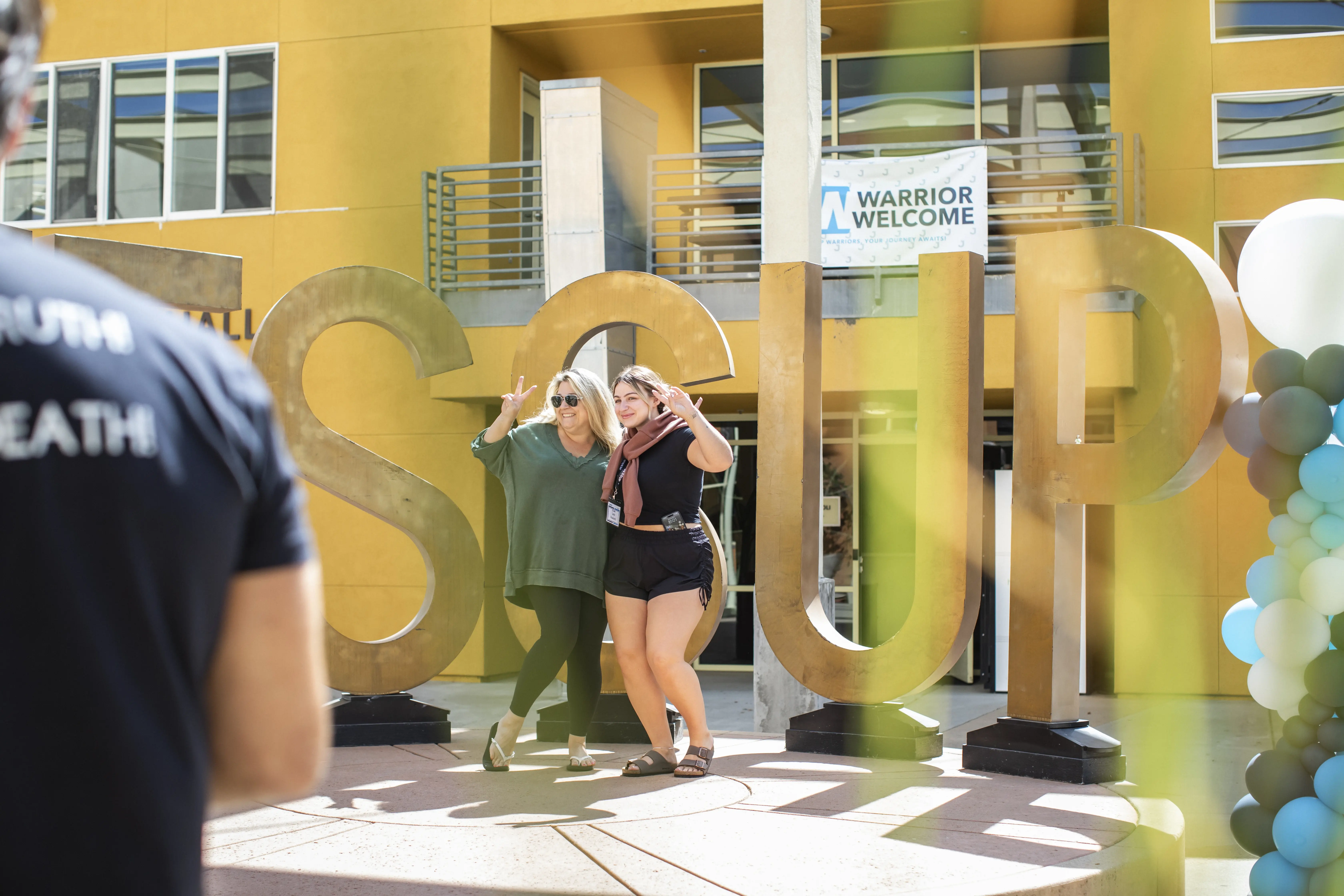 Two women posing and smiling in front of large gold letters spelling 'SOUP' outdoors with a yellow building and a 'Warrior Welcome' sign in the background.
