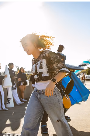 Young woman with curly hair wearing a black and white shirt and light blue ripped jeans walking outdoors with people and balloons in the background during daylight.