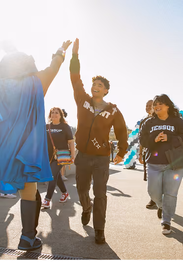 Young man smiling and giving a high five outdoors, surrounded by casually dressed people walking and chatting on a sunny day with balloons in the background.