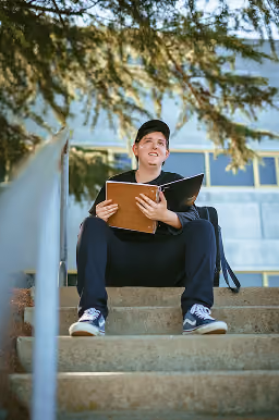 Young man wearing a black cap and dark clothes sitting on outdoor stone steps holding an open folder, with a backpack beside him and tree branches overhead.