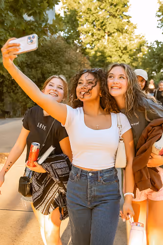 Three young women smiling and taking a selfie outdoors on a sunny day, surrounded by trees with other people in the background.