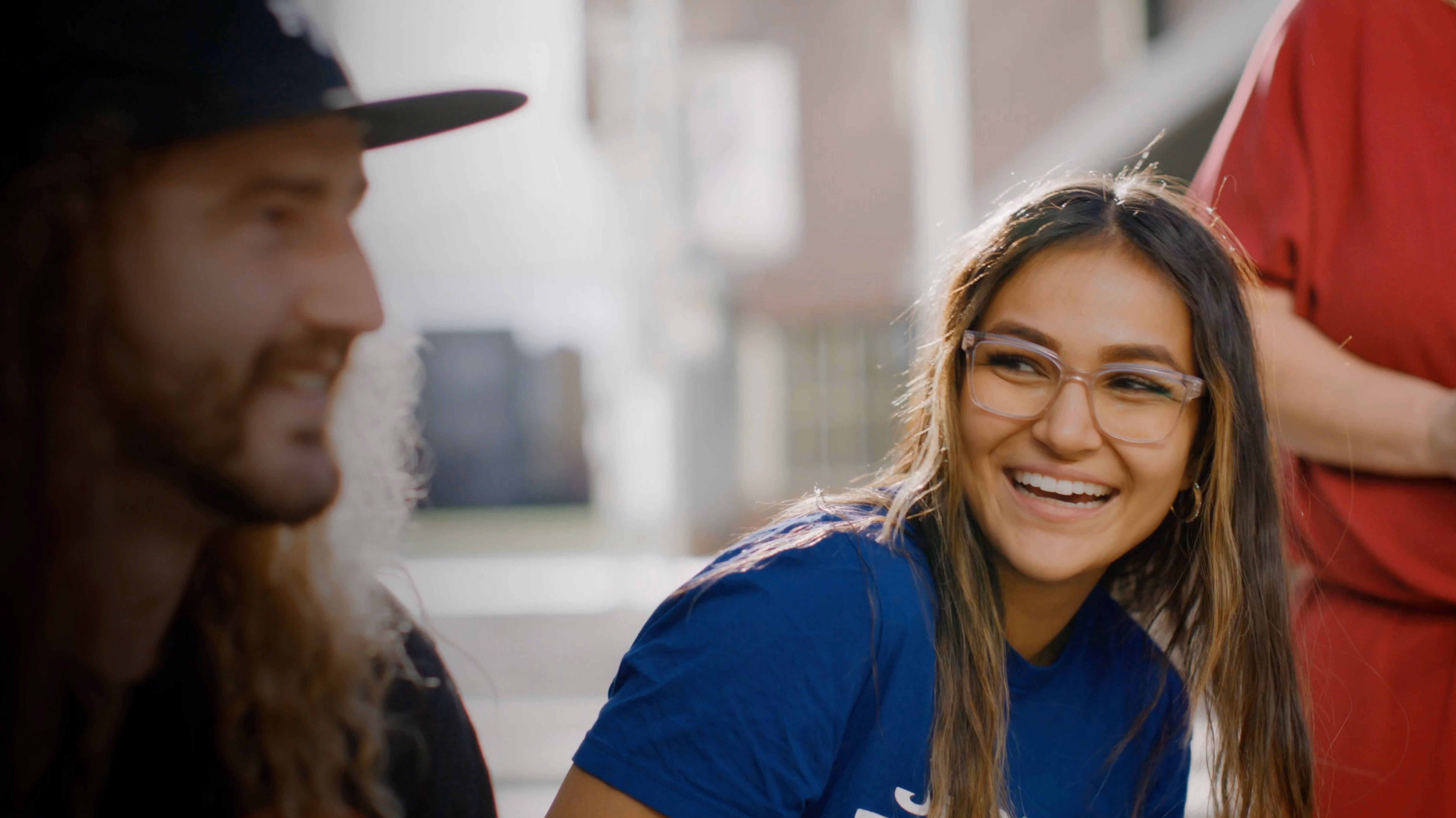 Smiling young woman with glasses and long hair wearing a blue shirt, looking at a man with long hair and a beard wearing a black hat.