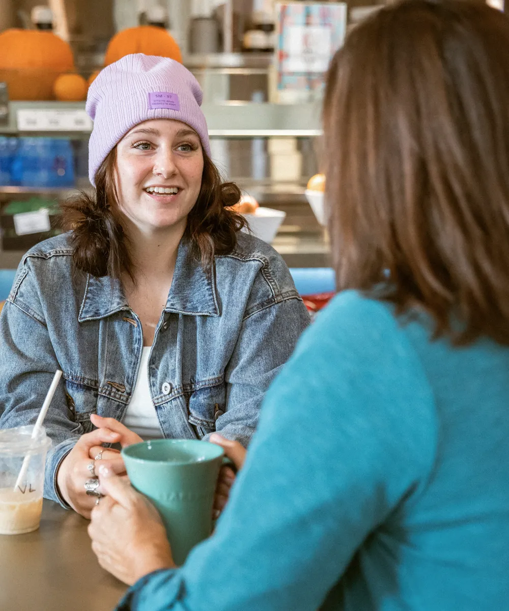 Two women sitting at a table in a cafe, one wearing a purple beanie and denim jacket, smiling and holding a drink while talking to the other woman in a teal top holding a green mug.