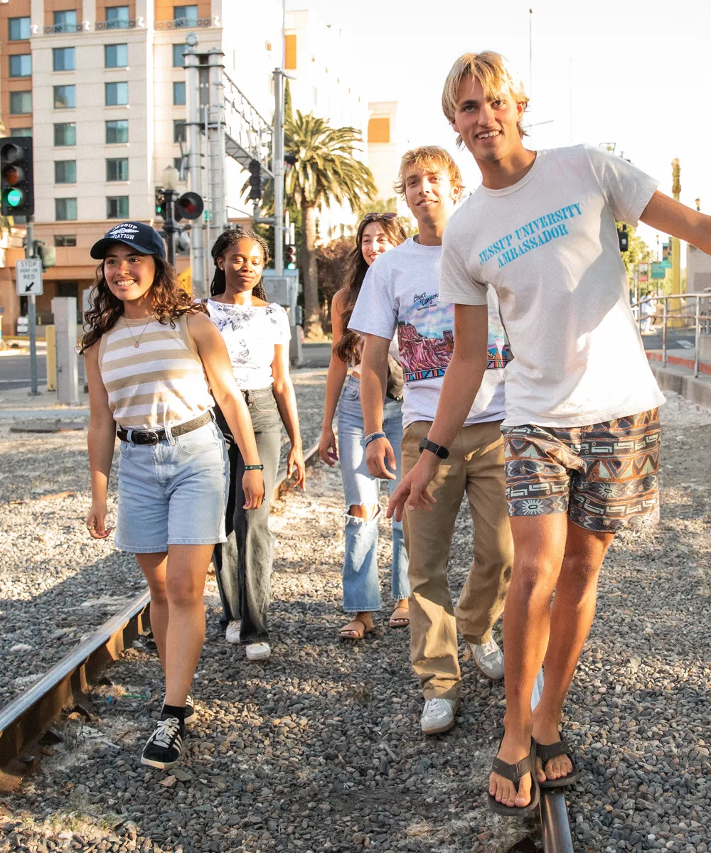 Five young adults walk along a gravel railroad track in an urban area with buildings and palm trees in the background; one person balances on the rail wearing a shirt that says Jessup University Ambassador.