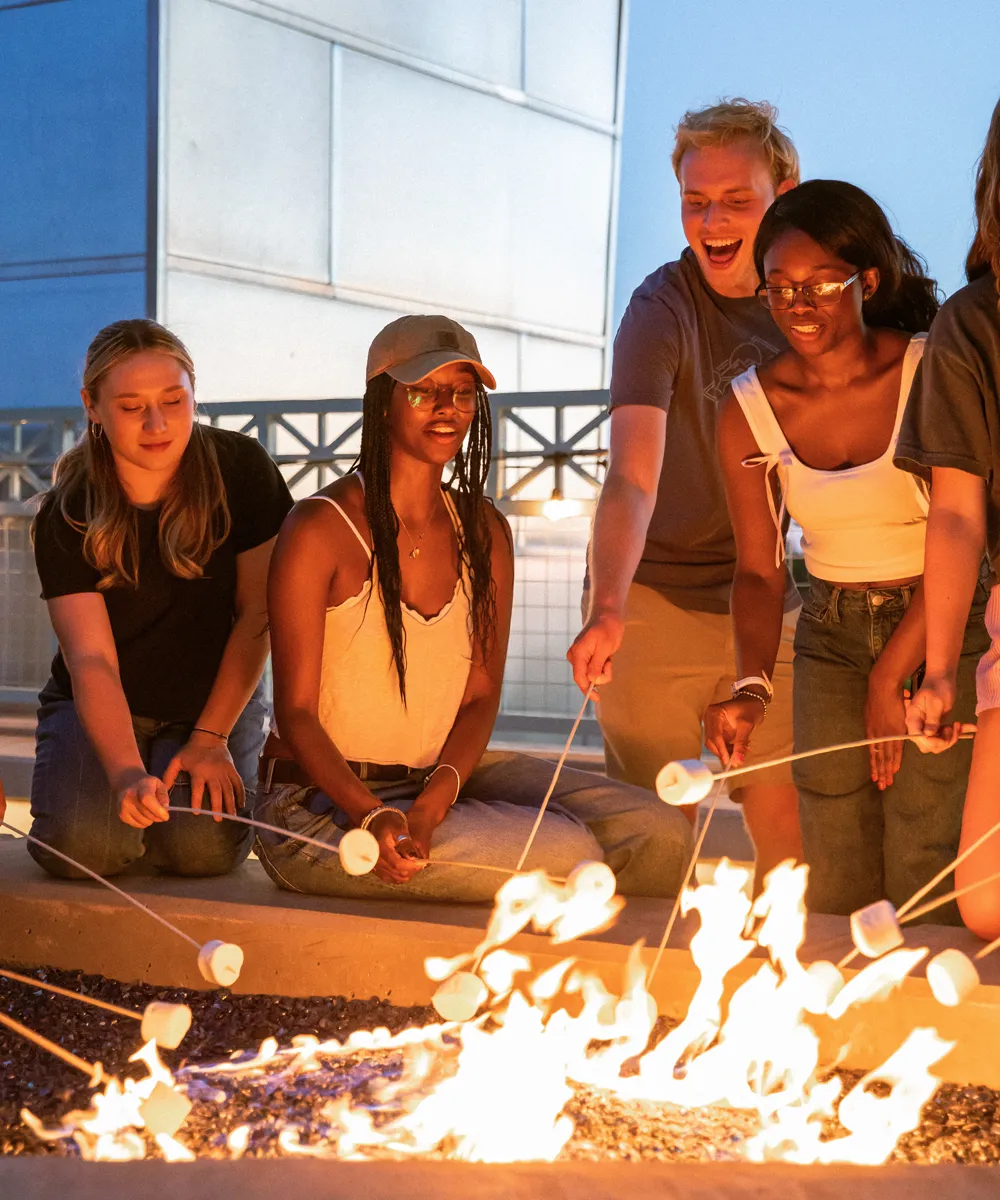 Group of young people around an outdoor fire pit roasting marshmallows on sticks during dusk.
