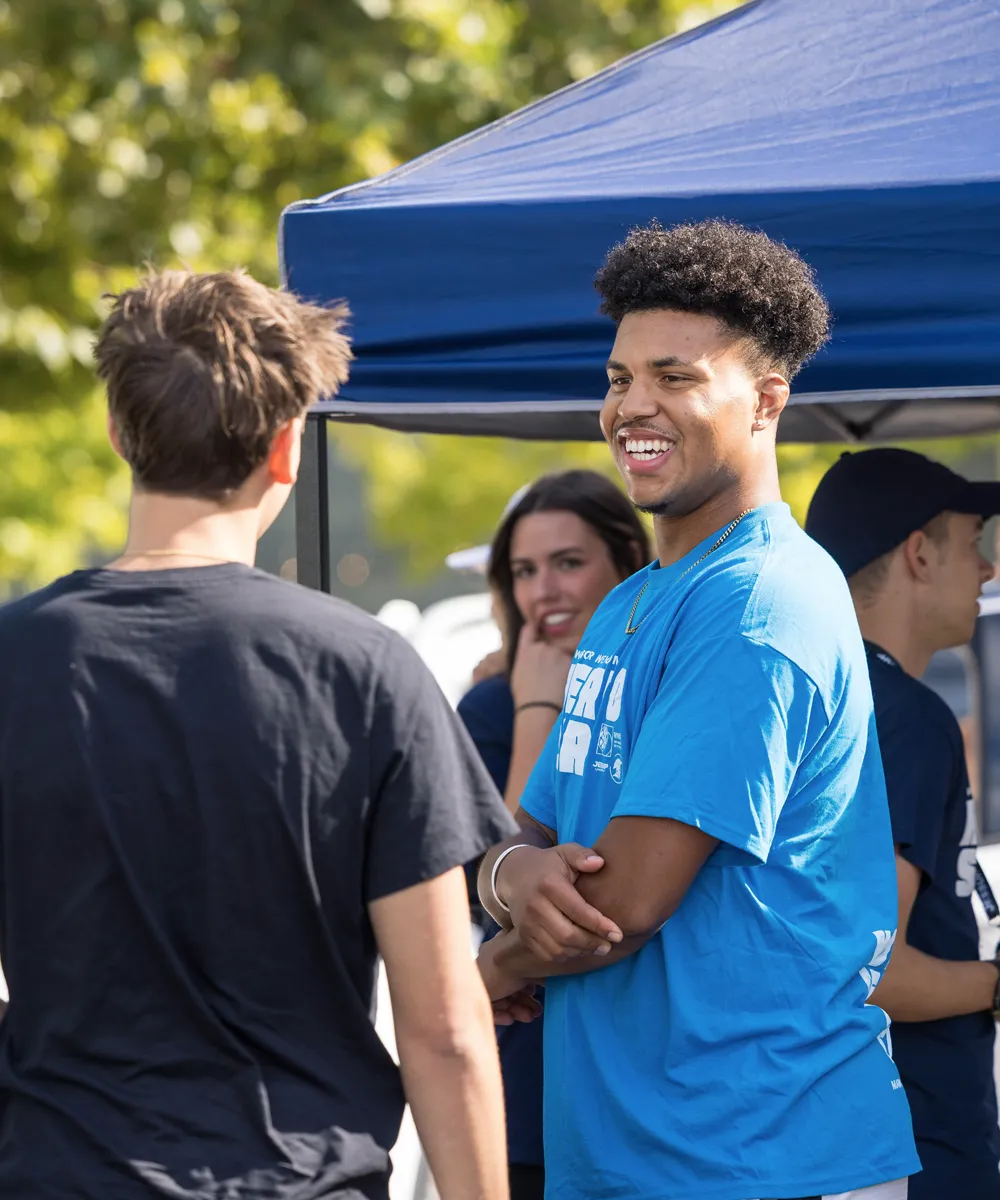 Group of young people talking and smiling outdoors near a blue canopy tent on a sunny day.