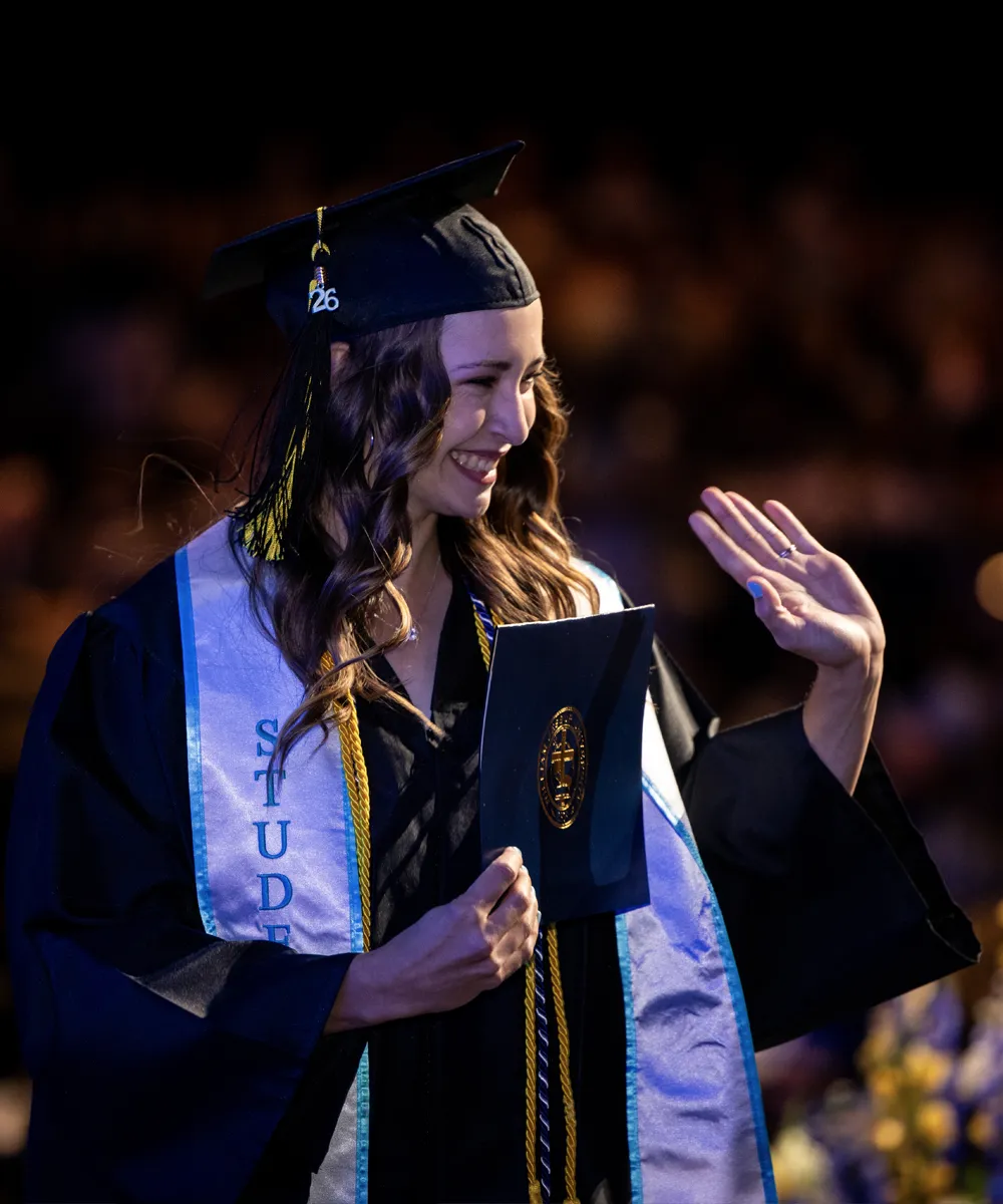 Smiling female graduate wearing a cap with tassel and gown, holding a diploma cover and waving with her left hand during a graduation ceremony.