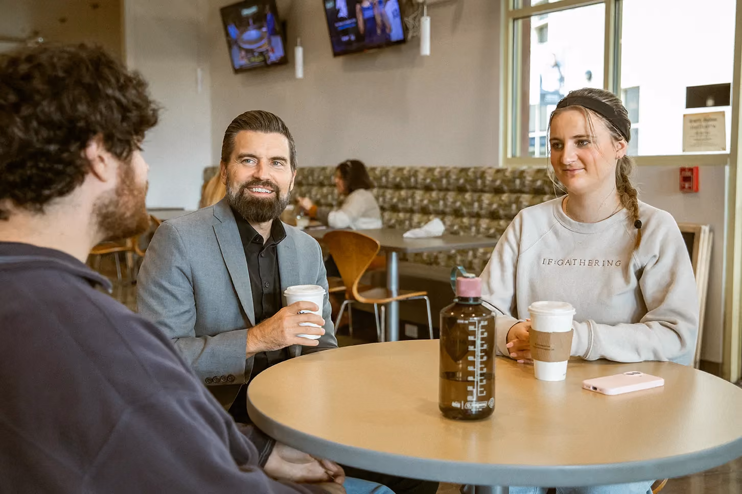 Three people sitting around a round table in a casual cafe setting, two holding takeaway coffee cups, one with a water bottle on the table, engaged in conversation with a relaxed atmosphere.