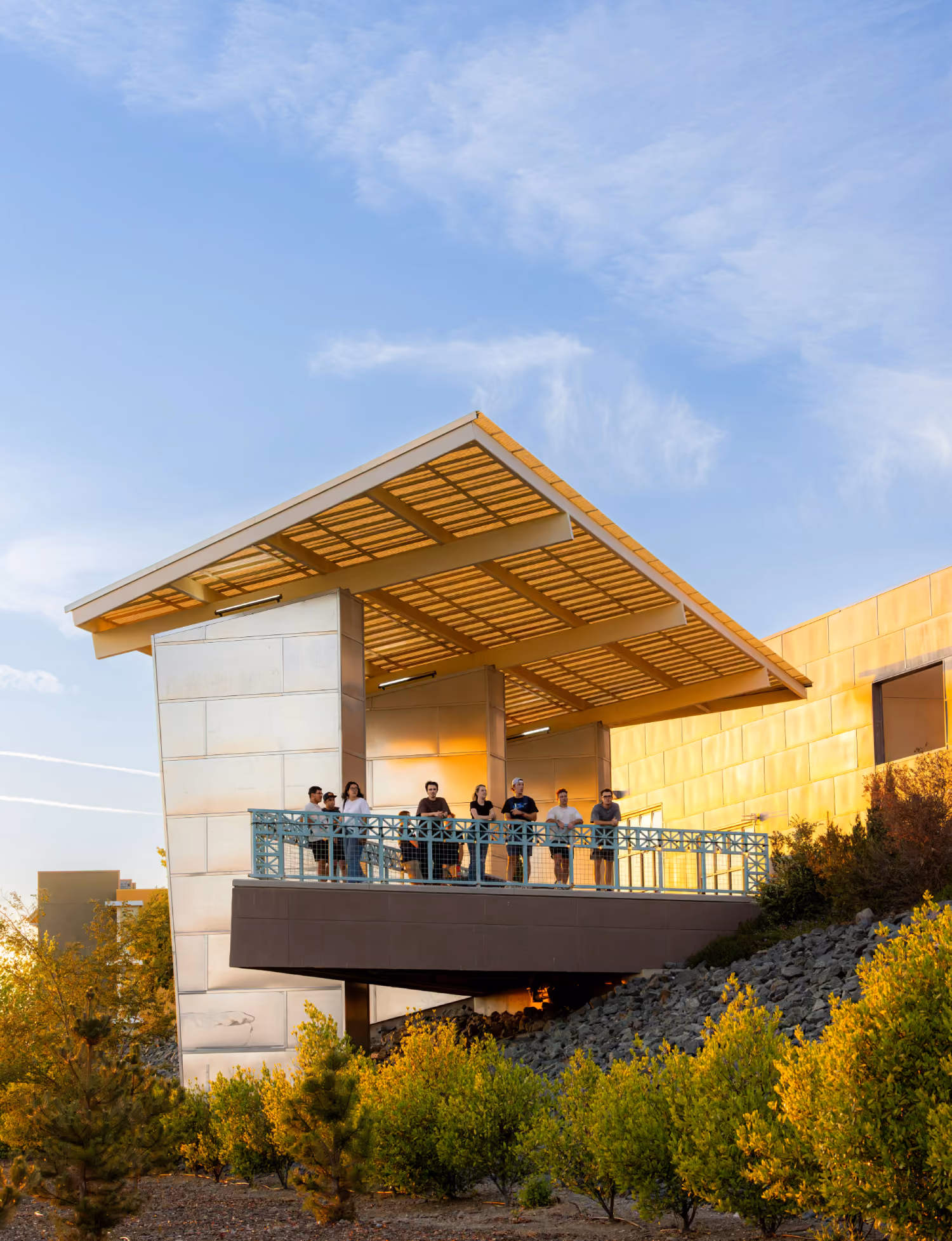 Group of people standing on an elevated balcony with blue railing under a large modern canopy on a sunny day with blue sky and green shrubbery below.