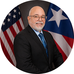 Official portrait of Lionel E. Santa Crispín in a suit with U.S. and Puerto Rico flags behind him.